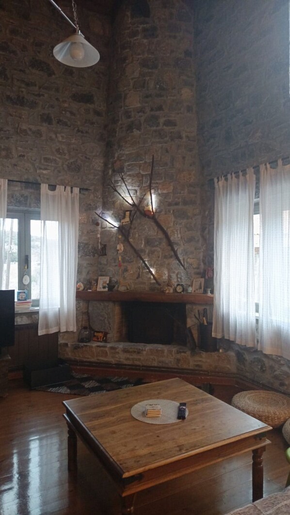 Une pièce rustique aux murs de pierre, dotée d’une cheminée, de meubles en bois et d’un luminaire suspendu.