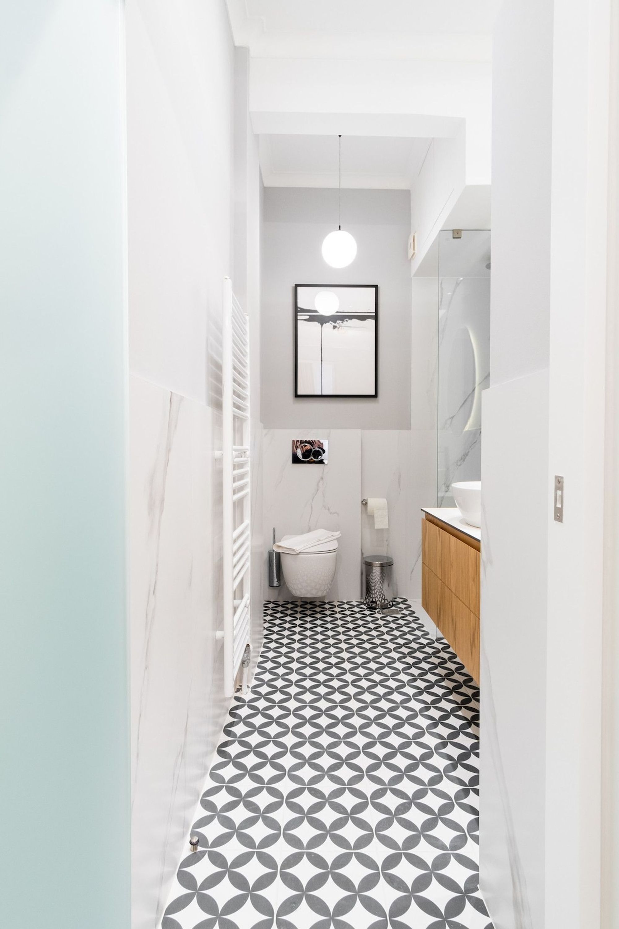 A narrow hallway with a geometric patterned floor, a white radiator, a hanging light fixture, a framed picture on the wall, and a wooden cabinet.