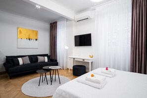 A modern hotel room with a black sofa, a round table, a bed with neatly folded white towels, a flat-screen TV, and a painting on the wall. - Kaplanon Residence Kolonaki (Athens)