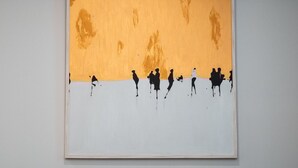 A framed artwork depicting silhouetted figures on a white background with an orange upper section, hung on a plain wall above a dark-colored sofa with two pillows. - Kaplanon Residence Kolonaki (Athens)