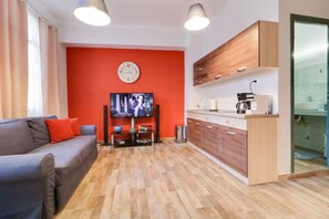 Una sala de estar moderna con un sofá gris, una pared de acento roja, un televisor de pantalla plana y una cocina de madera con armarios y una encimera.