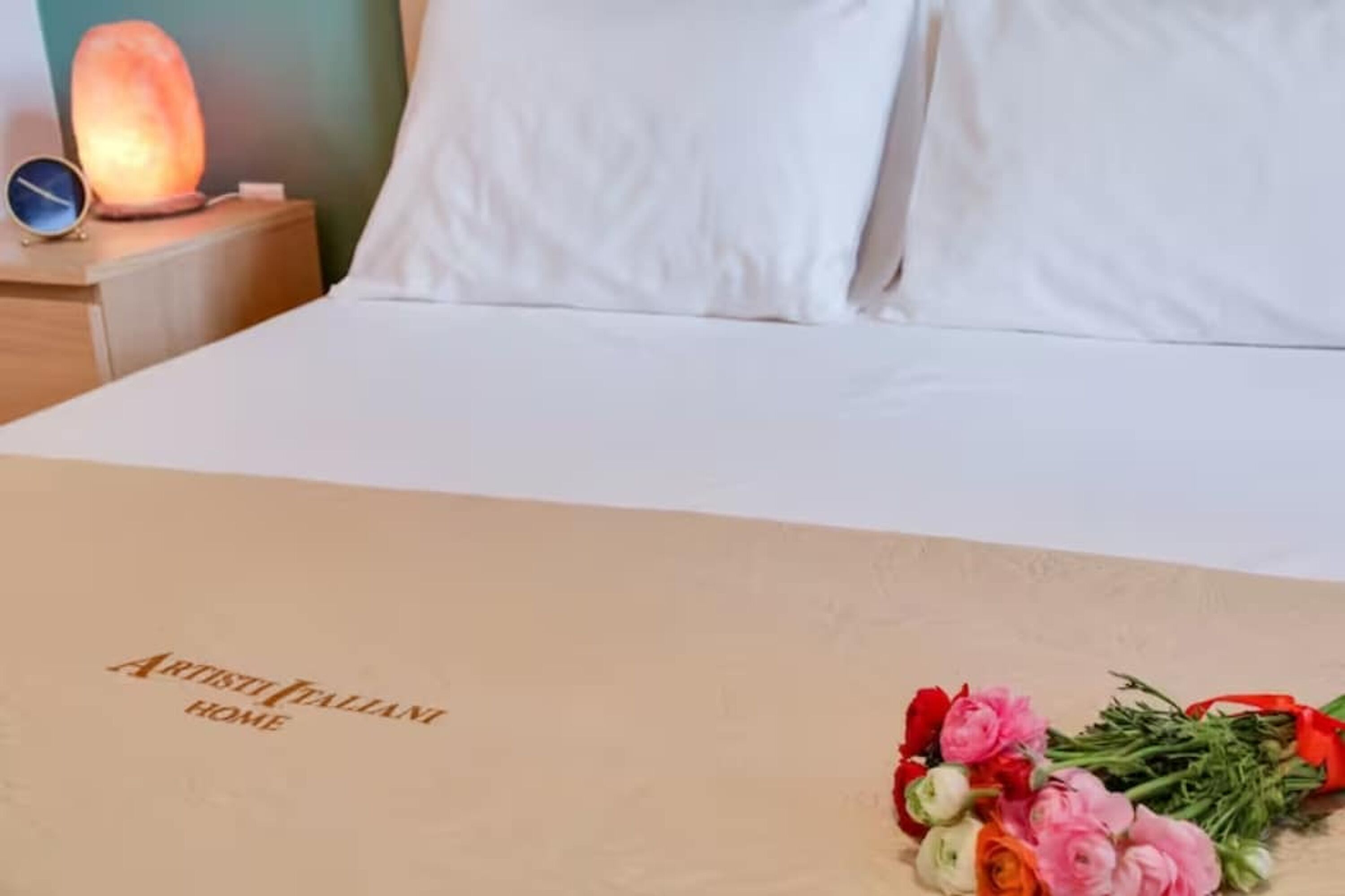 A neatly made bed with white linens, a bedside table with a salt lamp and a clock, and a card on the bed reading "ARTESI FALANI HOME".
