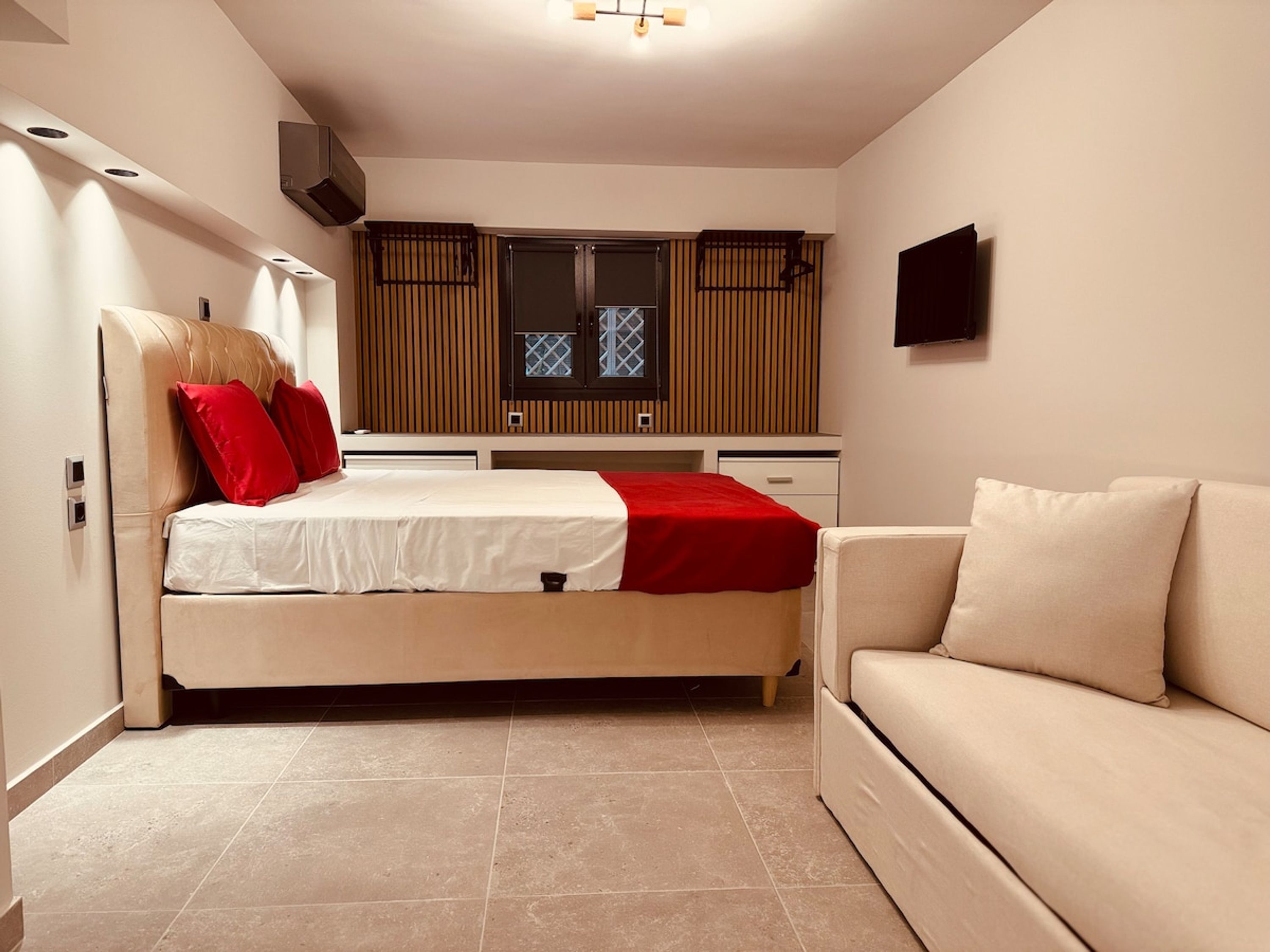 A compact bedroom with a bed, a sofa, a TV, and a window with blinds.