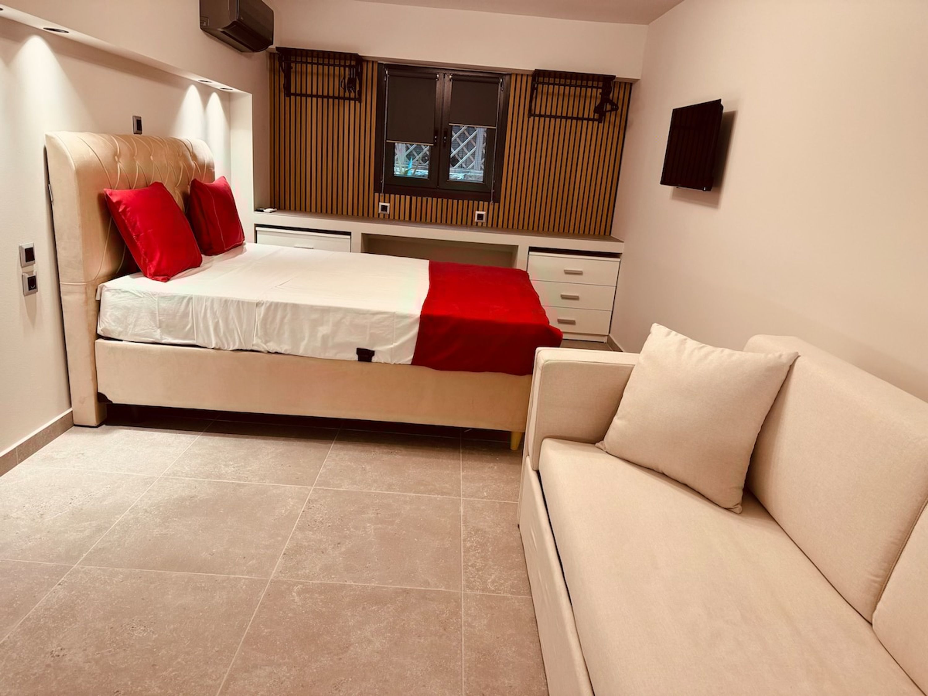 Une chambre à coucher avec un lit, une couverture rouge, un canapé beige et un téléviseur fixé au mur.