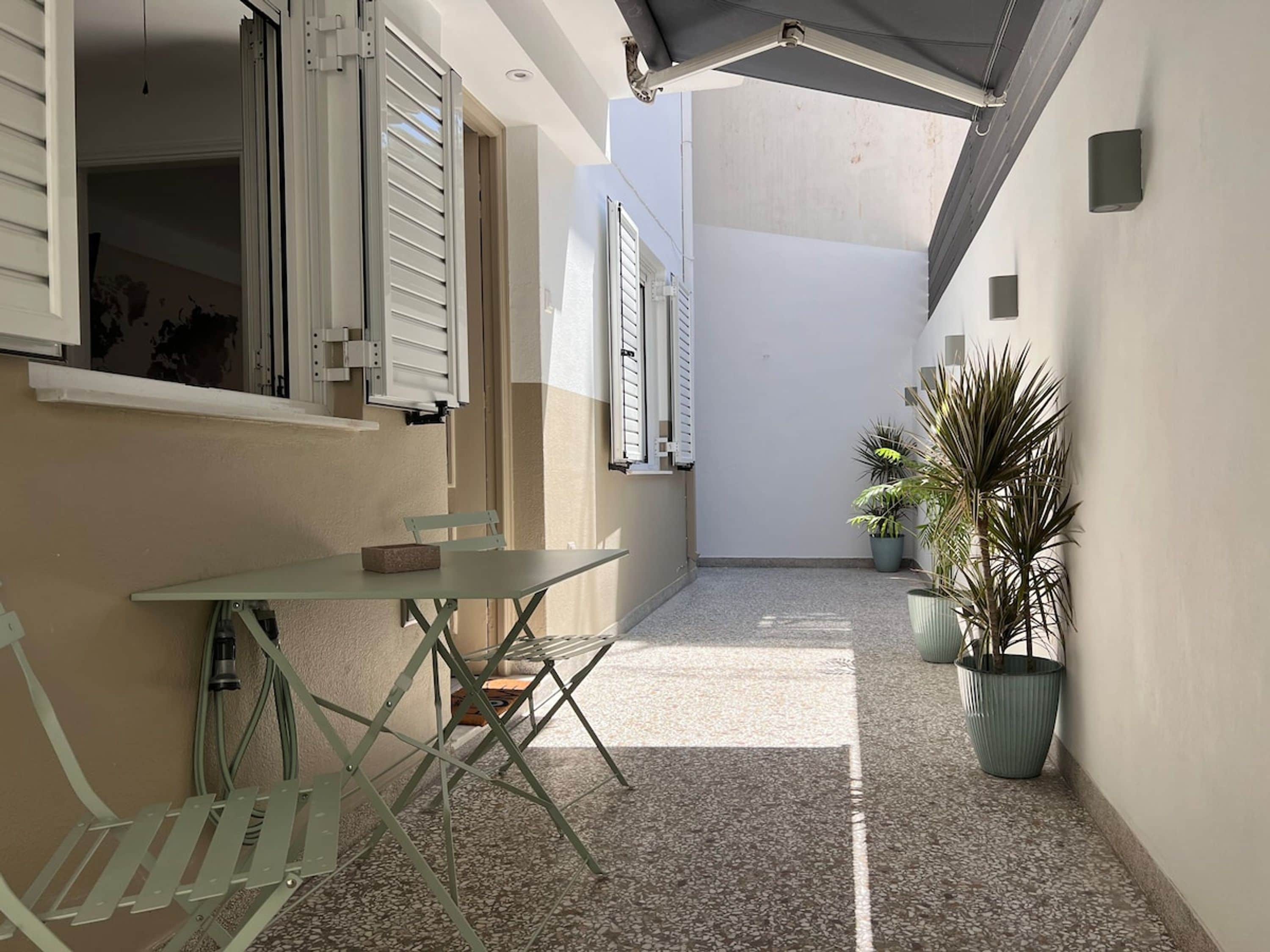 Um corredor externo estreito com uma mesa, cadeiras, plantas em vasos e uma porta que dá para um quarto.