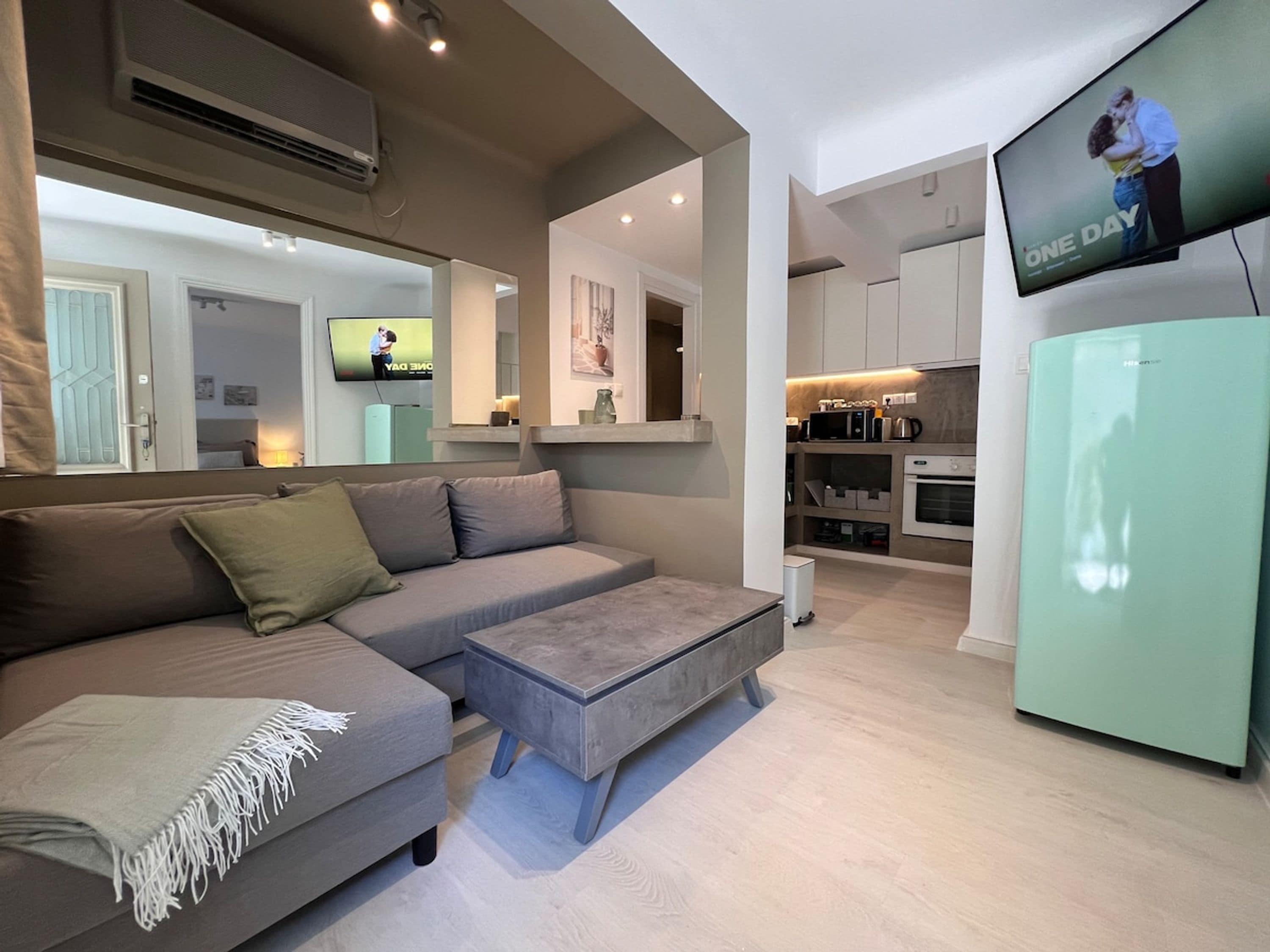 Ein modernes Wohnzimmer mit einem grauen Sofa, einem kleinen Hocker und einem an der Wand befestigten Fernseher.