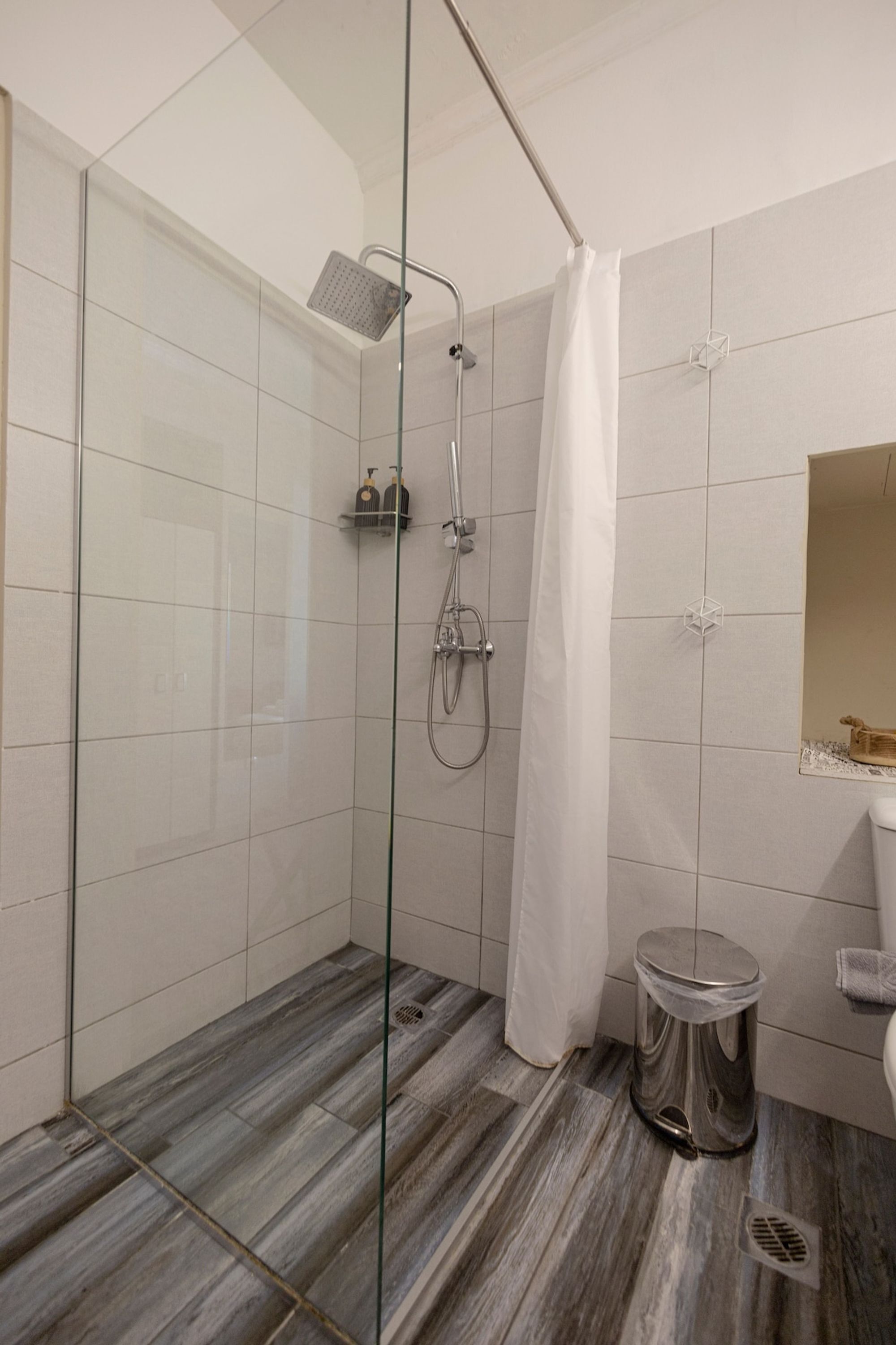 A modern bathroom with a glass shower enclosure, a marble floor, a toilet, and a small trash bin.