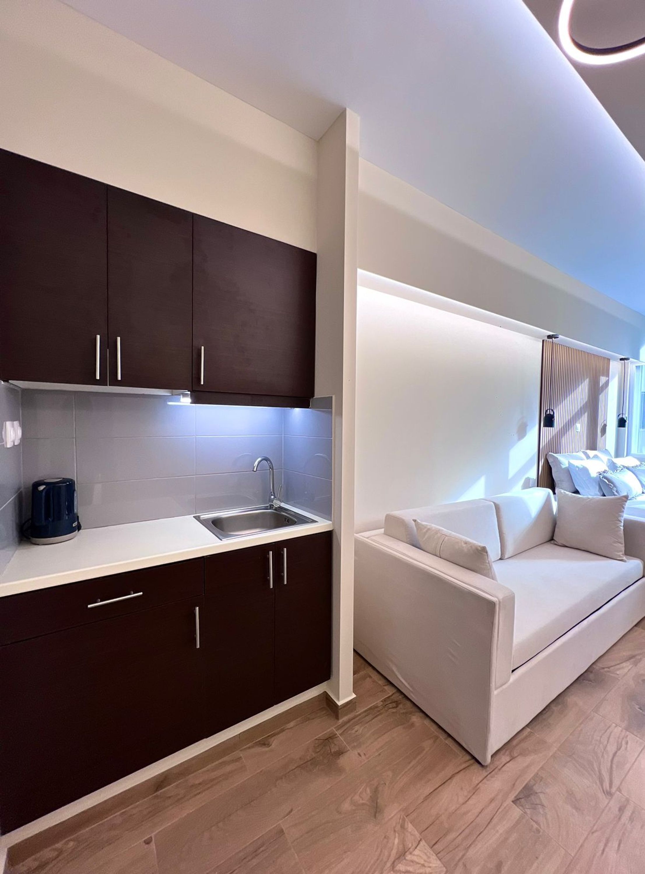 A modern kitchen with dark cabinets and a white countertop, a built-in sink, and a coffee maker. There is a white sofa with cushions in the adjacent living area.
