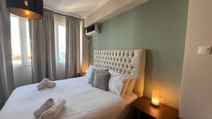 Un llit ben fet amb llençols blancs i dues tovalloles enrotllades, un capçal amb aspecte canelat, una taula de nit amb una làmpada, i una finestra amb cortines.