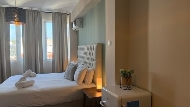Habitació d'hotel amb un llit gran, una taula de nit amb una làmpada, una petita planta i una finestra amb cortines.