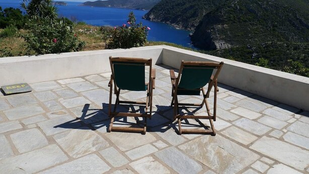 Two wooden chairs with green cushions on a stone patio overlooking a coastal landscape with a view of the sea and hills. - Gorgeous House Exclusive Location (Alonissos)