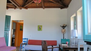 A cozy living room with a wooden ceiling, a red sofa, a wooden chair, a small table with a lamp, and a door leading to another room. - Gorgeous House Exclusive Location (Alonissos)