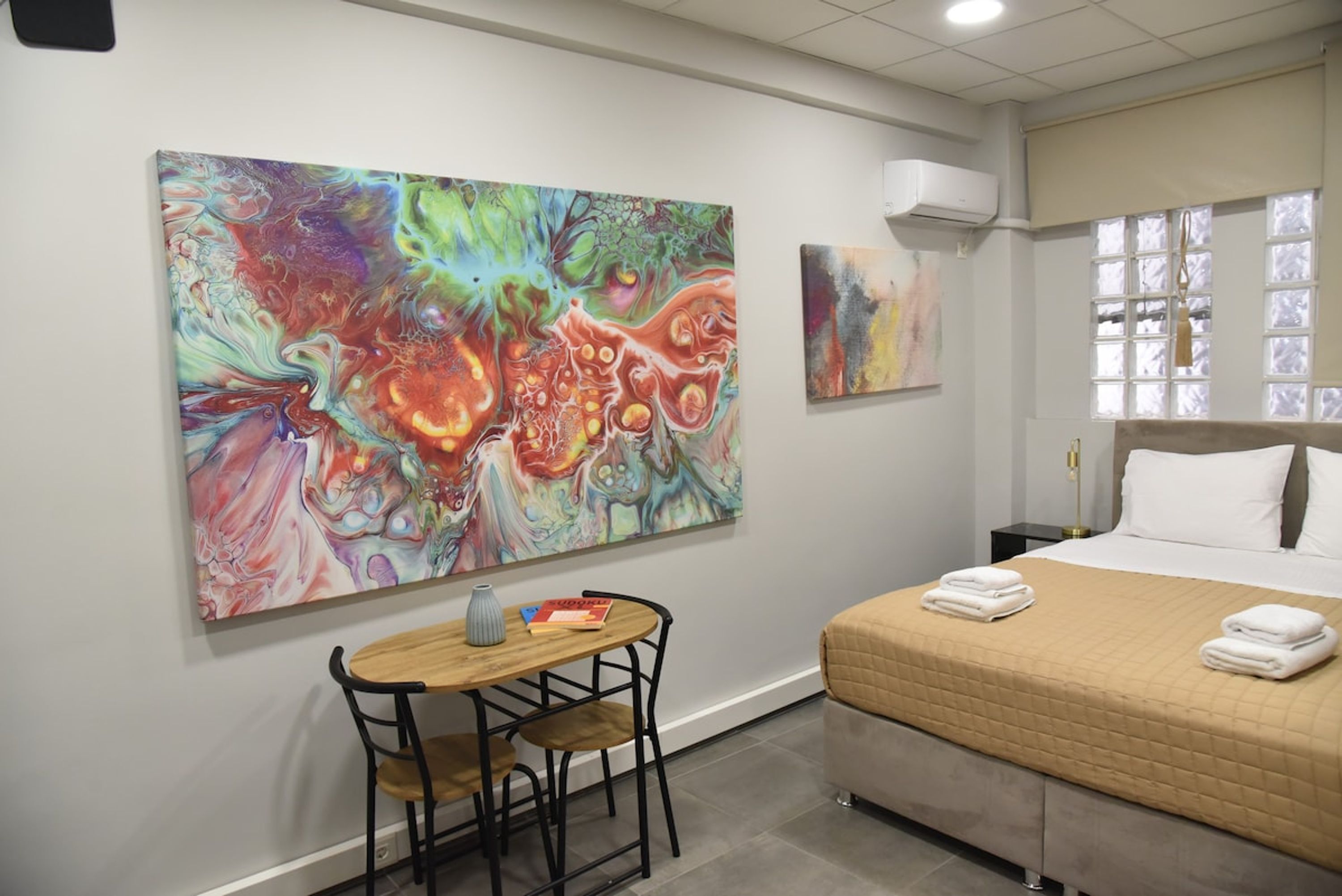 Habitación de hotel con un cuadro abstracto grande en la pared, una cama con colcha color crema y una mesita redonda con sillas.