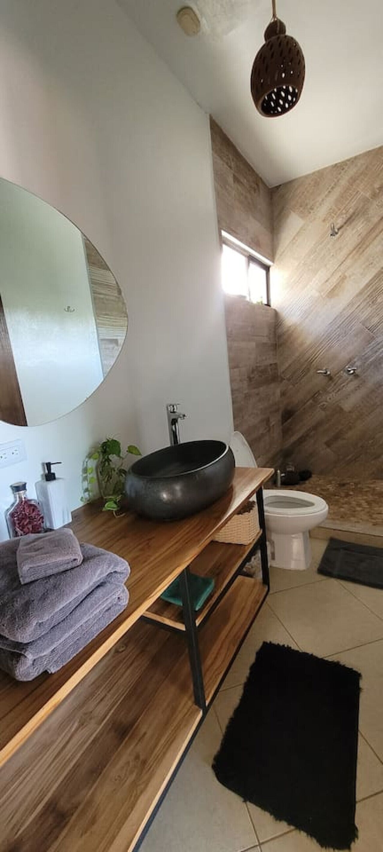 A modern bathroom with a round mirror, wooden vanity, black basin, and a large window.