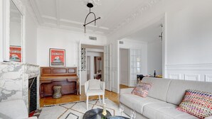 Ein großzügiger Wohnraum mit Marmorofen, einem Klavier, einer Couch mit bunten Kissen und einem Couchtisch mit dekorativer Ablage und Gläsern.