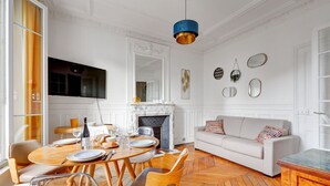 Un coin repas avec un parquet en bois disposé en chevrons, une table ronde dressée pour quatre personnes et un canapé blanc agrémenté de coussins à motifs.