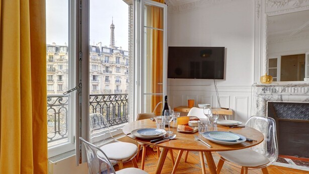 Un coin repas avec une table dressée pour quatre personnes, un téléviseur fixé au mur et une vue sur la Tour Eiffel, visible à travers de grandes fenêtres.