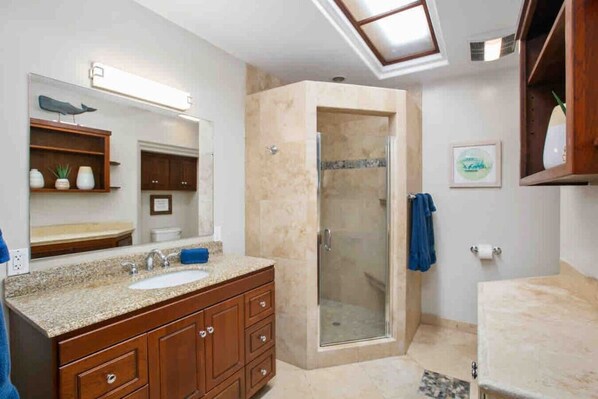 A bathroom with a granite countertop sink, wooden cabinets, a glass shower enclosure, and a skylight above the shower.