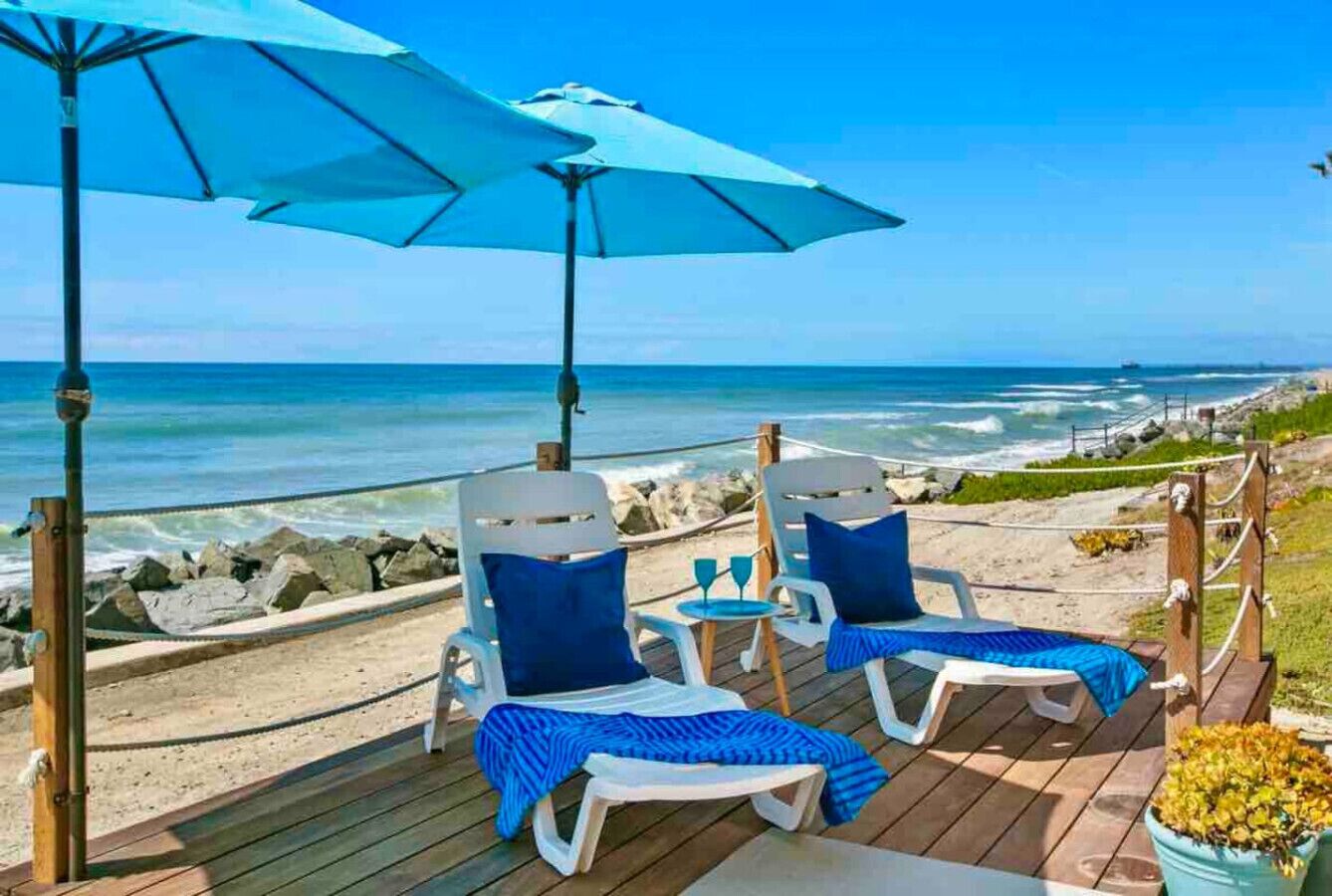 A beachside deck with two lounge chairs, a blue umbrella, and a view of the ocean.