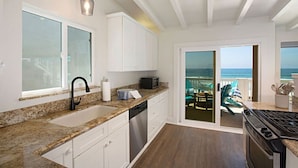 A kitchen with granite countertops, white cabinets, a dishwasher, and a stove. There is a large window and a door leading to a balcony with a view of the ocean.