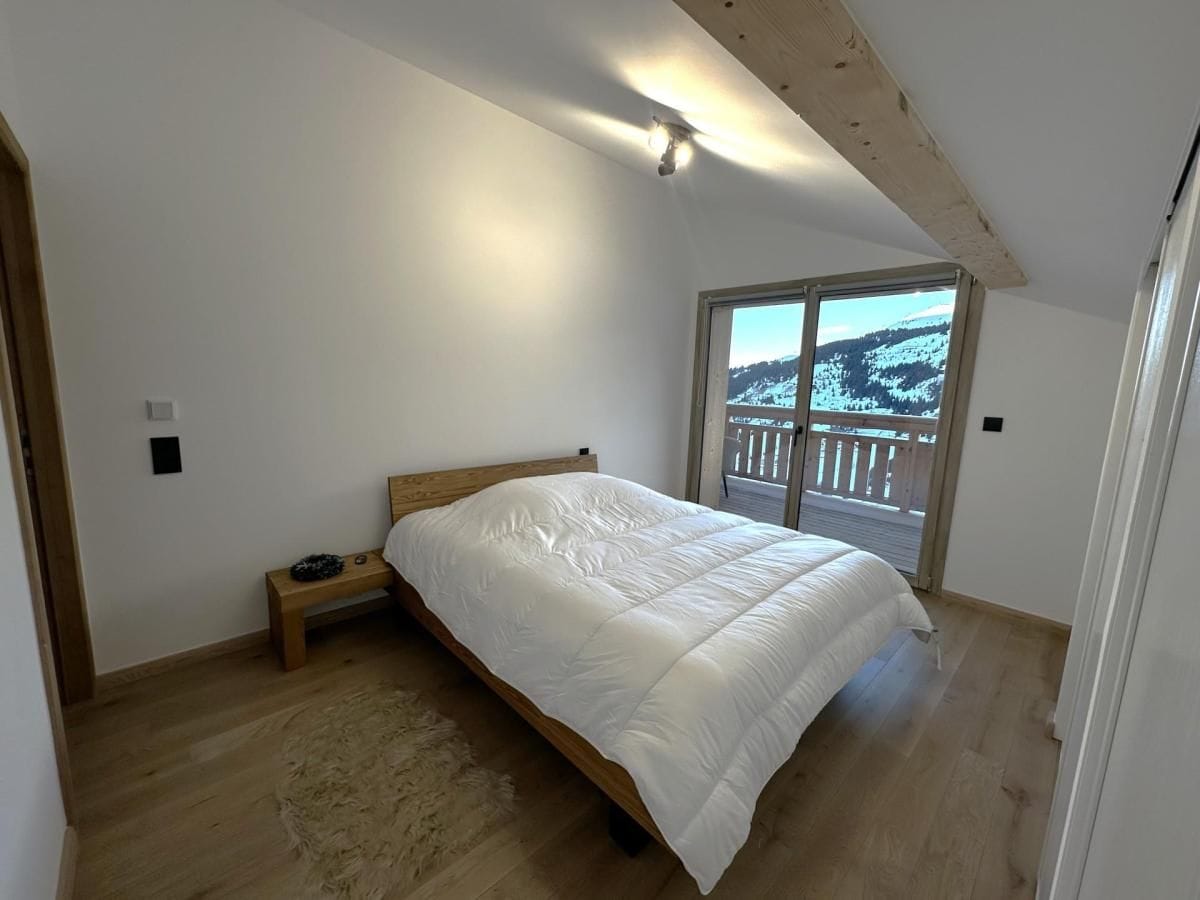 A bedroom with a wooden bed, a small bedside table, a large window with a view of a snowy landscape, and a wooden ceiling.