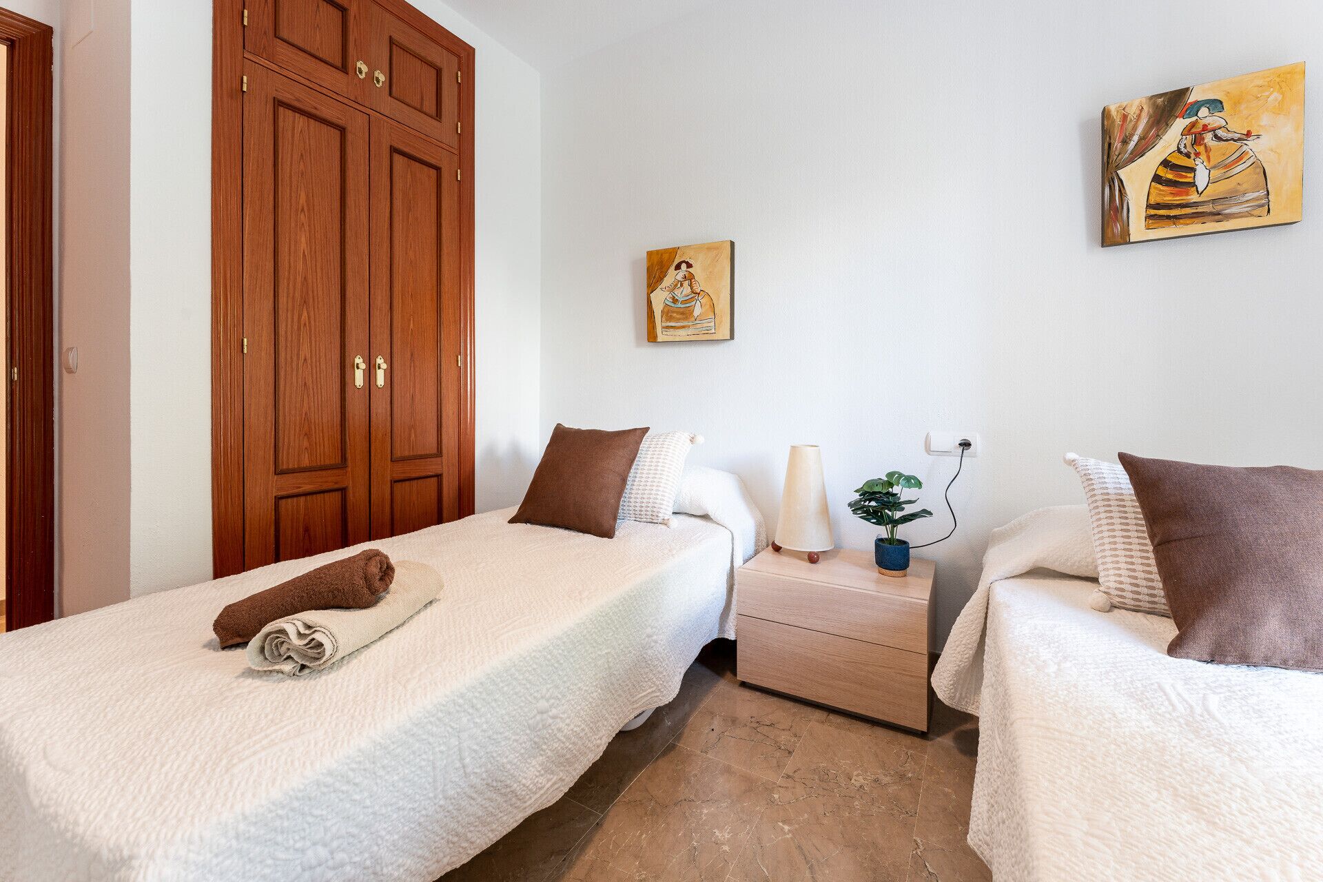 A bedroom with two single beds, a wooden wardrobe, a bedside table with a lamp, and a wall painting.