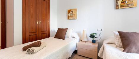 A bedroom with two single beds, a wooden wardrobe, a bedside table with a lamp, and a wall painting.