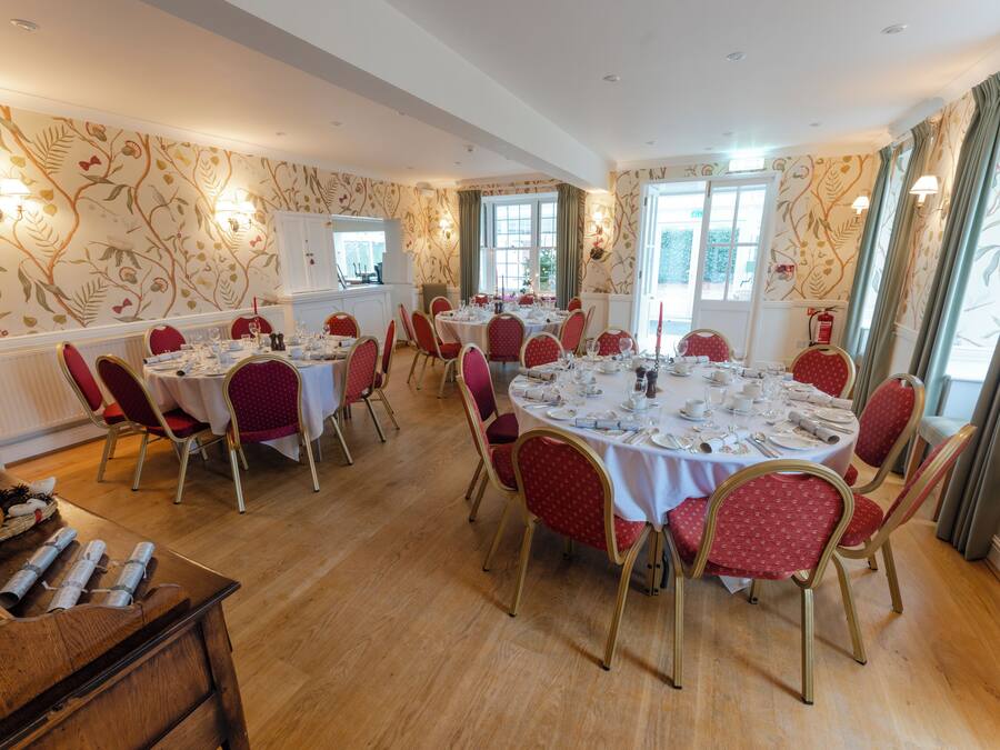 Uma sala de jantar com mesas redondas preparadas para vários convidados, cadeiras estofadas em vermelho e um aparador de madeira.