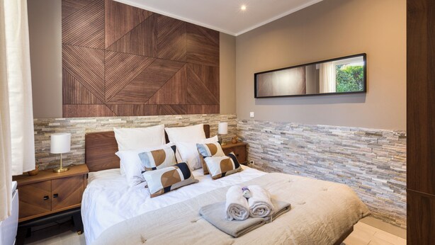 A modern hotel room with a large bed, a wooden headboard, a stone accent wall, a nightstand with a lamp, and a mirror on the wall.