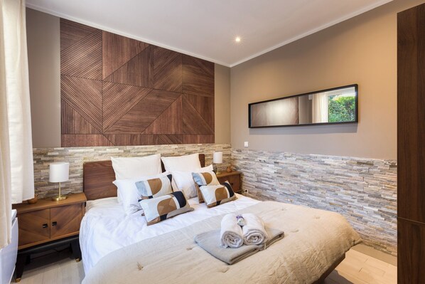 Quarto de hotel moderno com uma cama grande, cabeceira de madeira, parede de destaque em pedra, mesa de cabeceira com abajur e um espelho na parede.