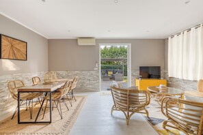 Ampla sala de estar com uma mesa de jantar, cadeiras de vime e um sofá. Há uma televisão e um armário amarelo ao fundo.