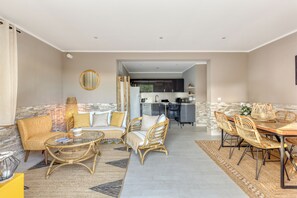 Ampla sala de estar com sofá, cadeiras, mesa de centro e mesa de jantar. Uma cozinha é visível ao fundo.