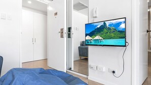 Makuuhuoneessa on sänky, seinälle asennettu televisio, jossa näkyy trooppinen maisema, vaatekaappi ja ovi.