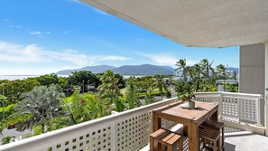 Un balcon avec une table et des chaises en bois, donnant sur un jardin luxuriant et offrant une vue sur l’océan, avec des montagnes à l’horizon.