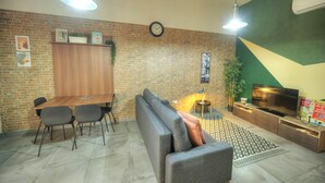 Un salon moderne avec un mur en briques, une table à manger avec des chaises, un canapé, un téléviseur et une horloge.
