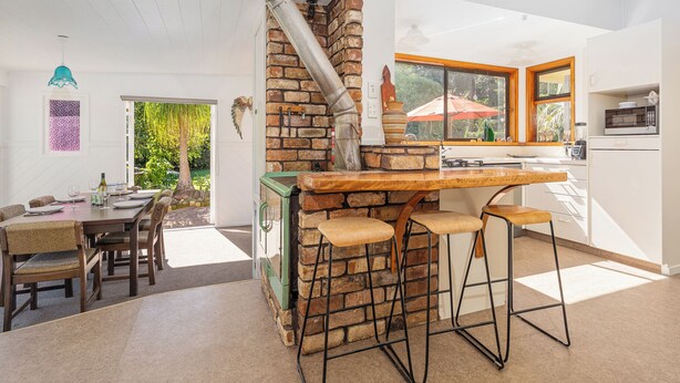 A kitchen with a brick stove, wooden bar stools, and a dining area with a table and chairs. - Birdsong - Whangamata Holiday Home (Whangamata)