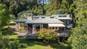 A two-story house with a deck and a covered porch, surrounded by lush greenery and trees. - Birdsong - Whangamata Holiday Home (Whangamata)