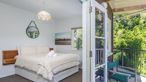 A bedroom with a bed, a bedside table, a hanging light fixture, a window, and a view of greenery outside. - Birdsong - Whangamata Holiday Home (Whangamata)