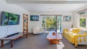 A spacious living room with a large window, a wooden coffee table, a sofa, a chair, and a TV mounted on the wall. - Birdsong - Whangamata Holiday Home (Whangamata)