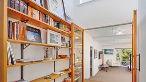 A well-organized library with bookshelves, a reading area with a chair and table, and a view of the outdoors through a glass door. - Birdsong - Whangamata Holiday Home (Whangamata)
