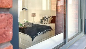 A bedroom with a large window, a bed with a black bedspread, and a brick wall visible through the window.