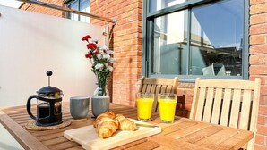 A wooden table with croissants, a French press, and two glasses of orange juice, set against a brick wall with a window and a vase of flowers.