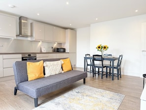 A modern kitchen with white cabinets, a grey sofa with yellow cushions, and a dining table with chairs. - South-Facing Balcony Apt, Canterbury City Centre (Canterbury)