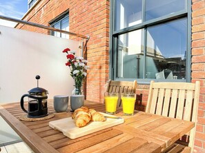 A wooden table with croissants, a coffee pot, two glasses of orange juice, and a vase with flowers.