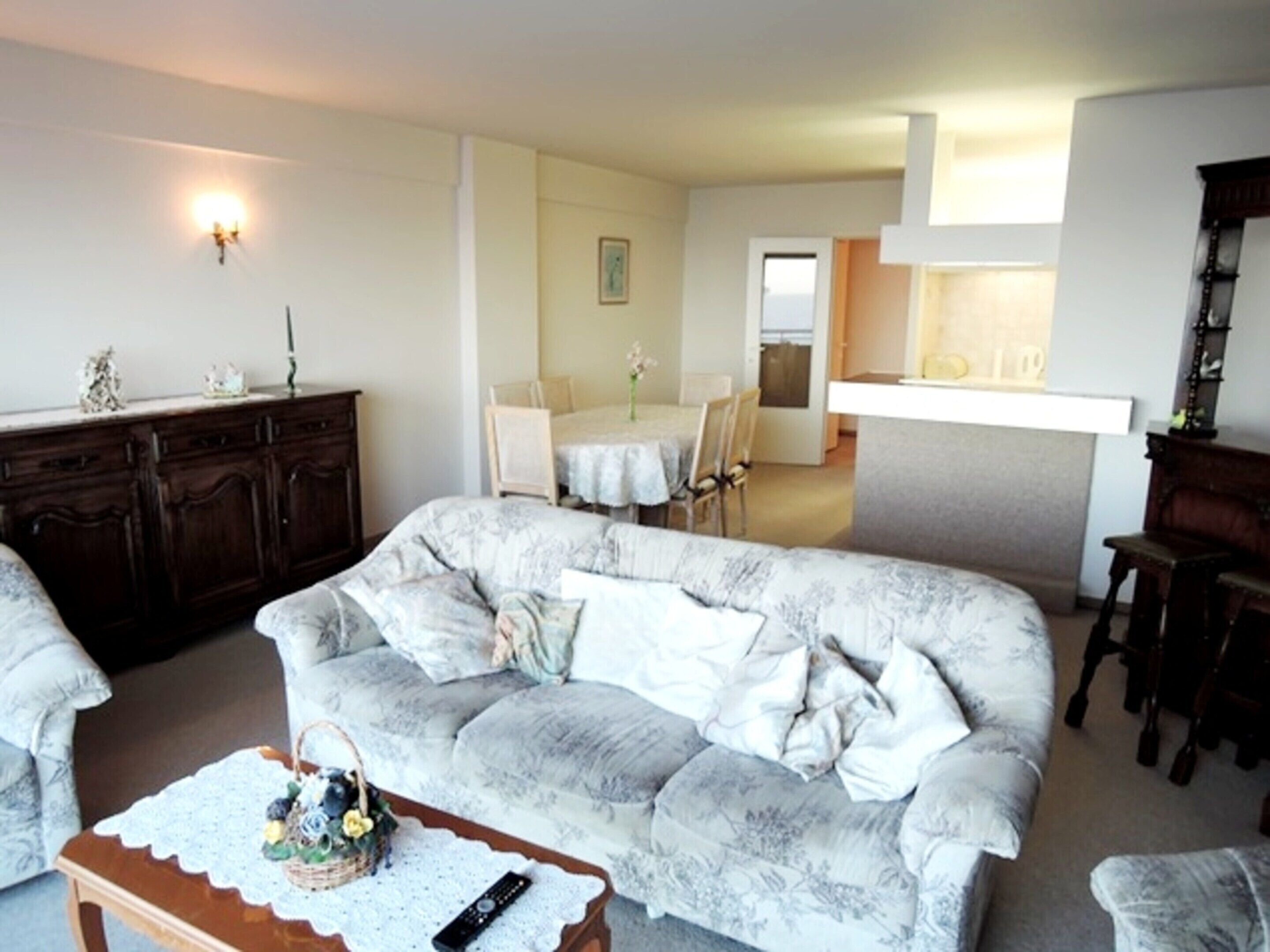 A living room with a sofa, a coffee table with a basket and remote, a dining area with a table and chairs, and a kitchen area with a counter and cabinets.