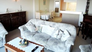 Un salon comprenant un canapé, une table basse avec un panier et une télécommande, un coin repas avec une table et des chaises, et une cuisine avec un comptoir et des placards.