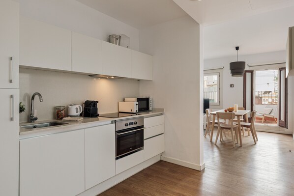 A modern kitchen with white cabinets, a microwave, a kettle, and an oven. There is a dining area with a table and chairs. - OLA LIVING ST ANTONI 17 (Barcelona)