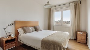 A neatly arranged bedroom with a wooden headboard, a bed with a beige blanket, a bedside table with a lamp, and a view of a cityscape through a window with curtains. - OLA LIVING ST ANTONI 17 (Barcelona)