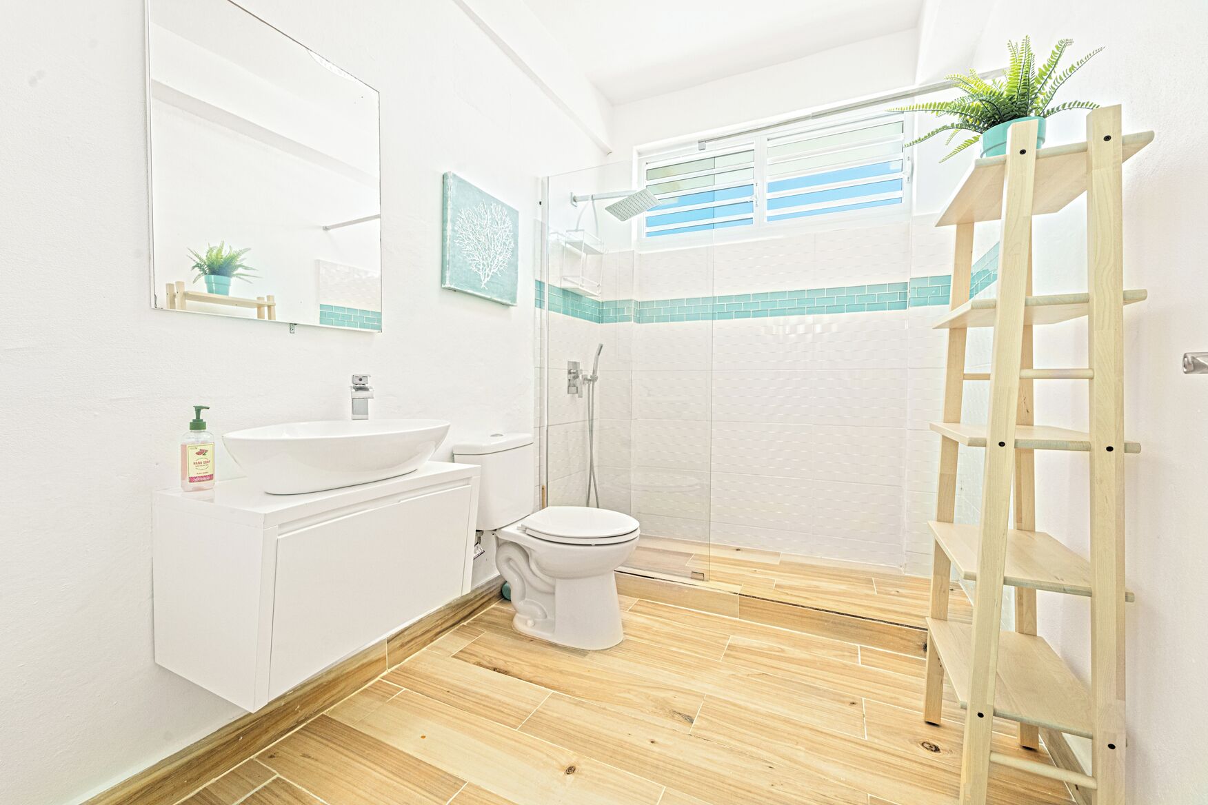 Un baño con lavabo, inodoro y ducha blancos. Hay un estante de madera con una planta encima.