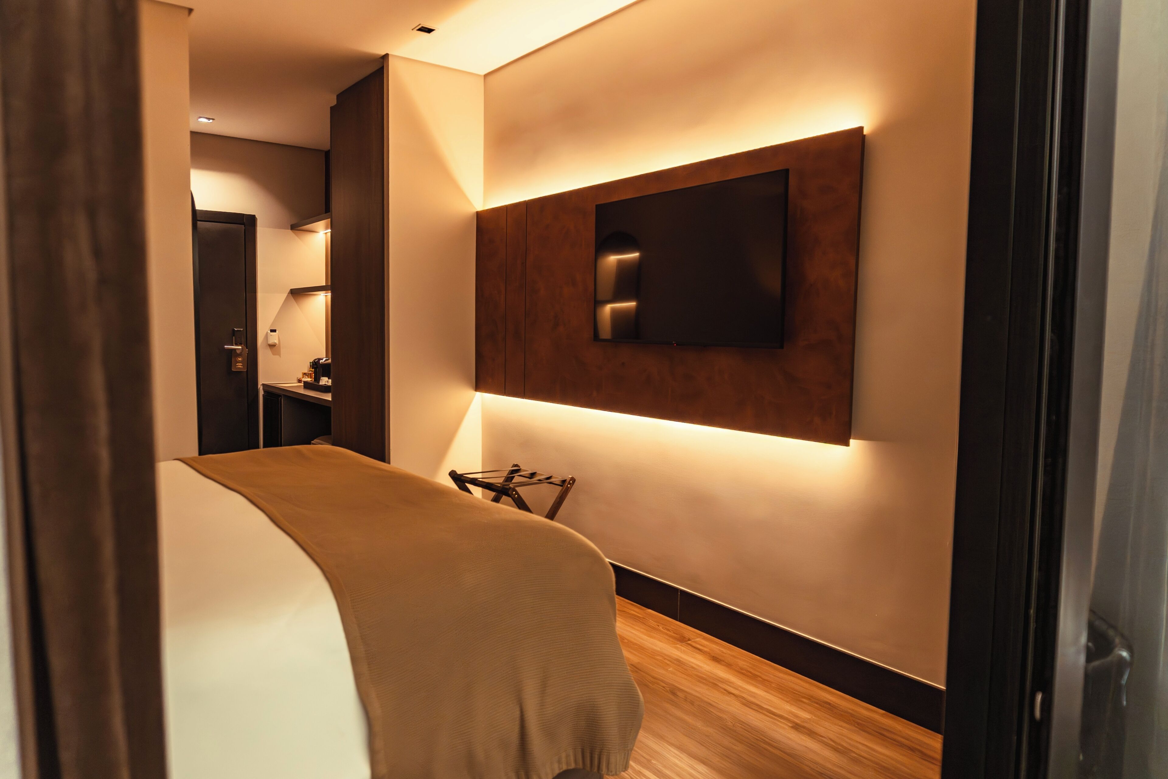 Un pasillo moderno de hotel con piso de madera, una cama con manta color beige, un televisor montado y una puerta que da a otra habitación.